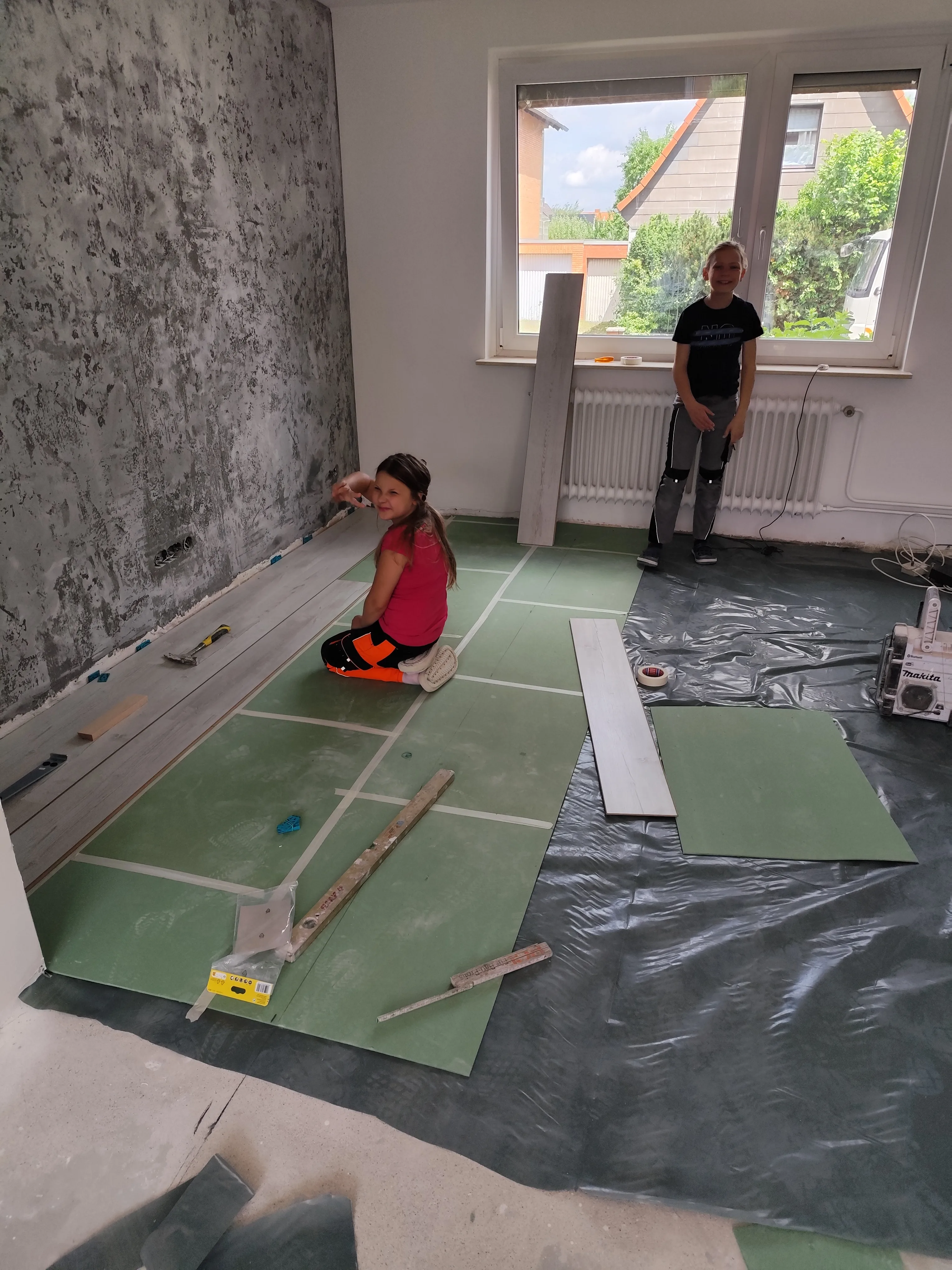 Ein Mädchen verlegt Bodenbelag in einem Raum, während ein Junge im Hintergrund zuschaut