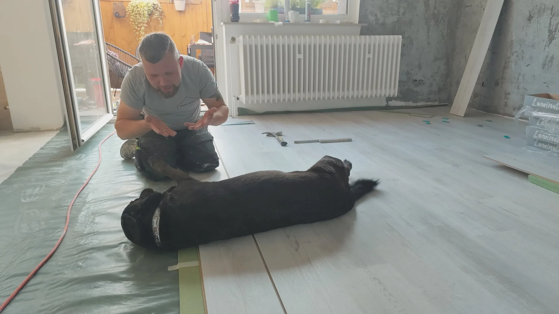 Facharbeiter verlegt einen Bodenbelag, während ein Hund entspannt auf dem Boden liegt